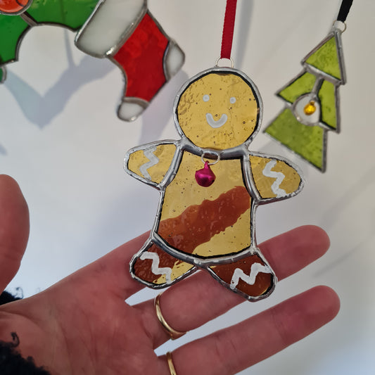 a staine glass ornament of a gingerbread man. amber muffled glass with white paint icing detail and a pink bell.