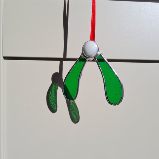 a stained glass mistletoe ornament made wihta medium green glass and a white glass nugget. hanging on a red ribbon.