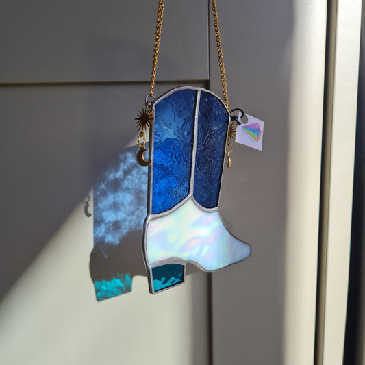 a blue and white stained glass cowboy boot suncatcher. the blue glass has a patterned texture. the boot hangs on a gold chain with star and moon gold charms.