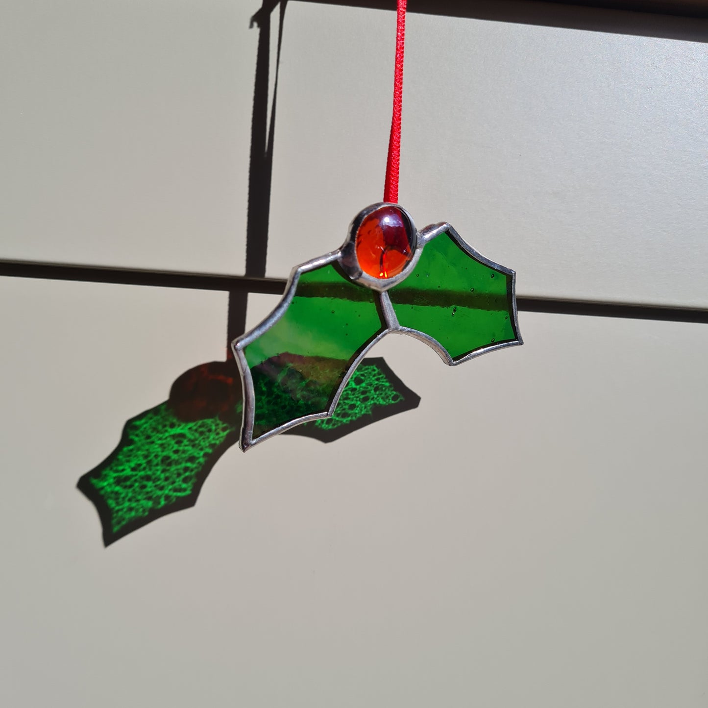 small stained glass hanging ornament, with two green glass holly leaves and a red glass nugget joining them. 